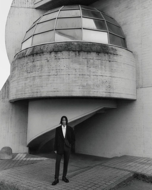 Monochrome shot of a male model in a suit posing in front of a modern architectural building featuring geometric shapes and a domed structure.