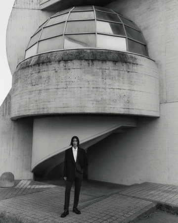 Monochrome shot of a male model in a suit posing in front of a modern architectural building featuring geometric shapes and a domed structure.