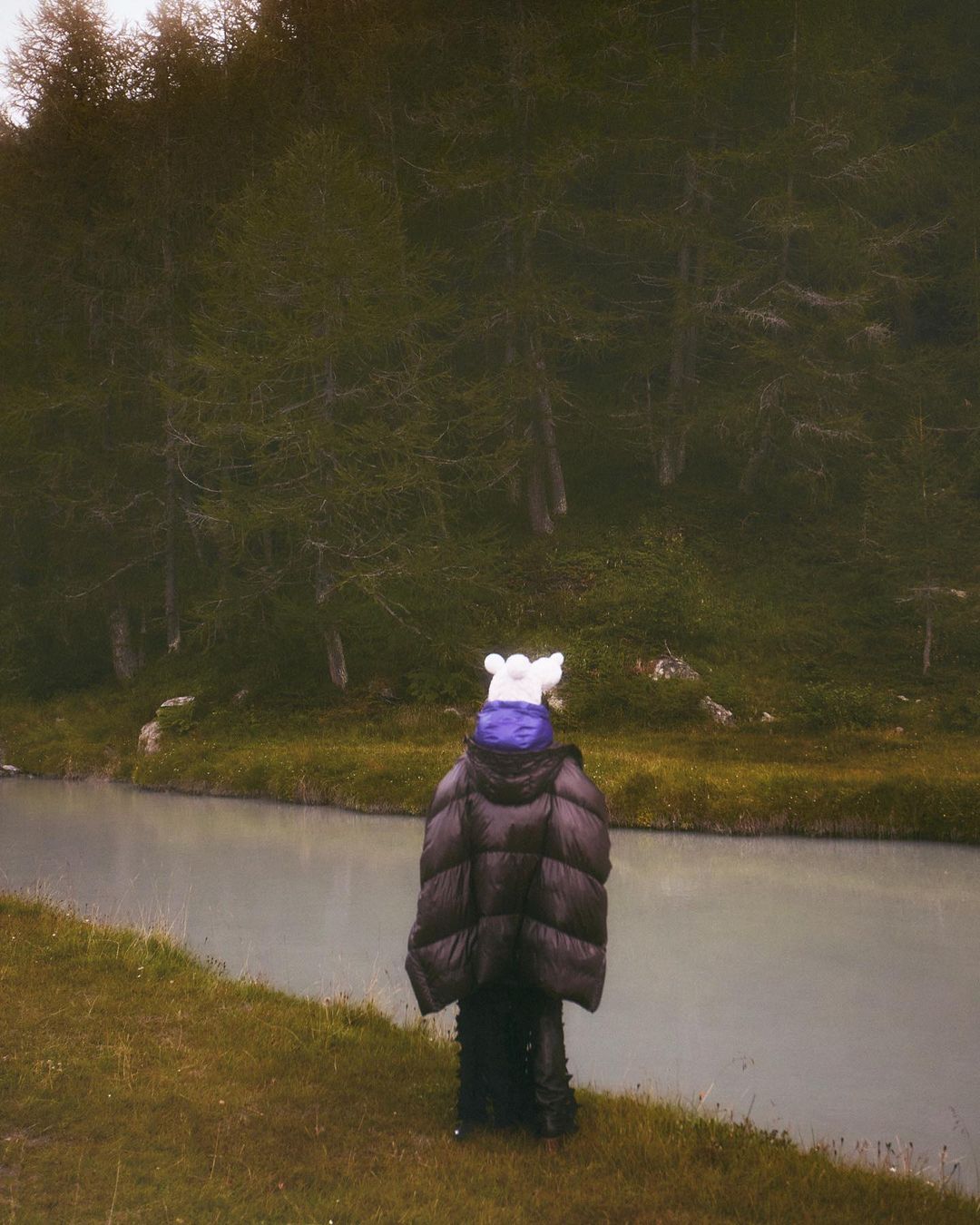 Person in oversized puffer jacket and animal hat stands by blue-gray river with forest background on an overcast day in nature.