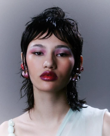 Close-up portrait of a model with dark wet hair, pink eyeshadow, glossy red lips, and sculptural earrings against a grey background.