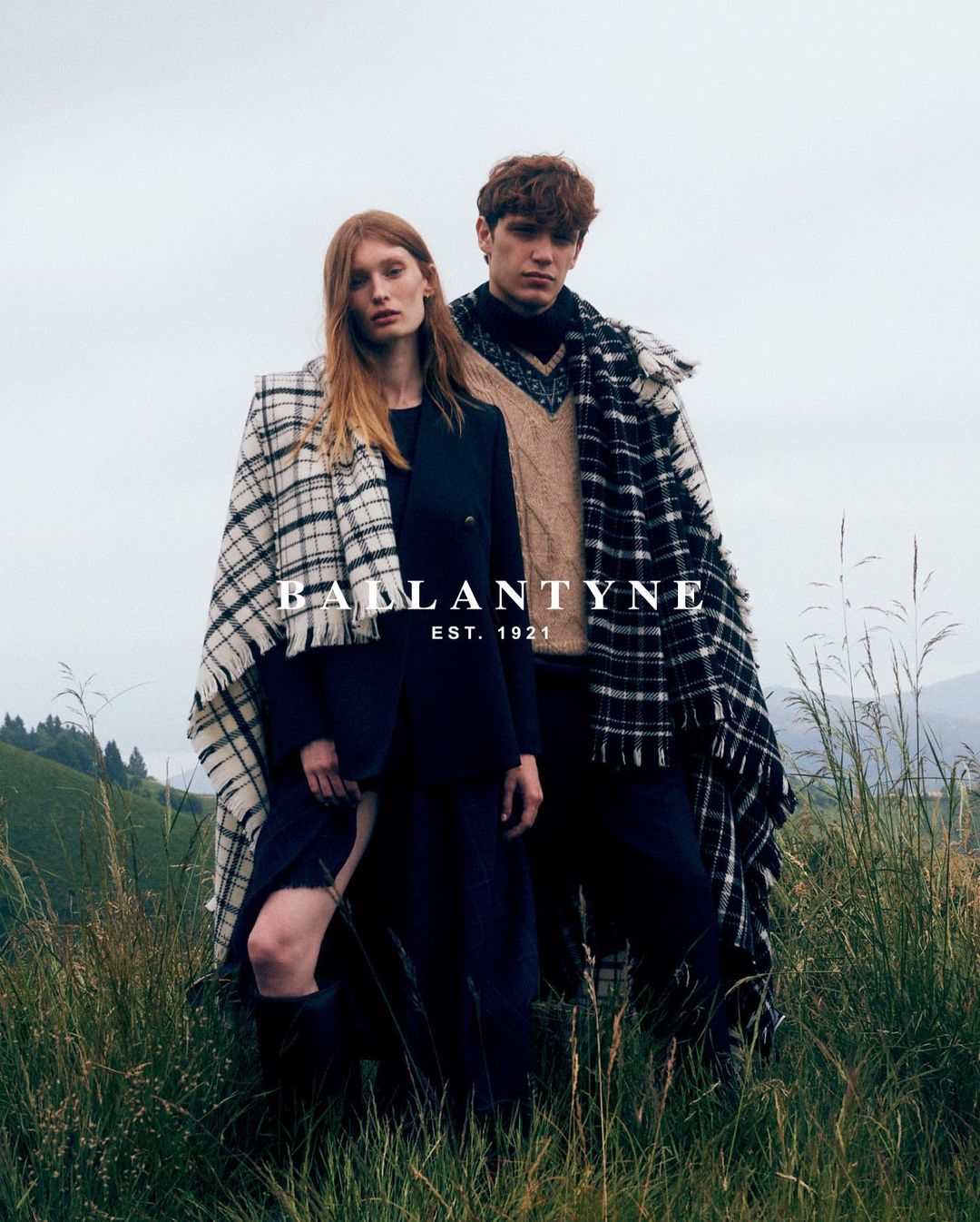 Two fashion models in outdoor setting wearing shawls and dark clothing with long grass. Brand is Ballantyne Est. 1921.