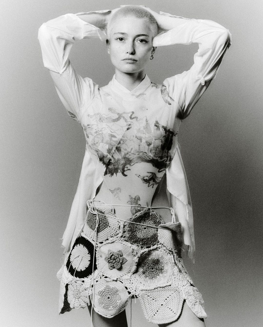Young woman in a light shirt and crochet skirt poses in a studio with hands behind her head, showcasing fashion and style.