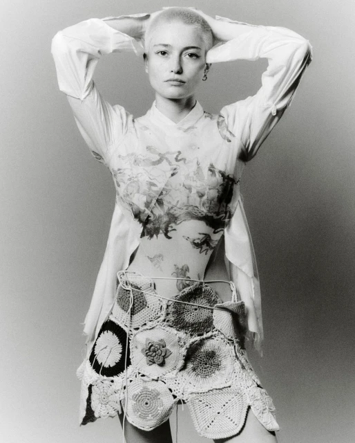 Young woman in a light shirt and crochet skirt poses in a studio with hands behind her head, showcasing fashion and style.