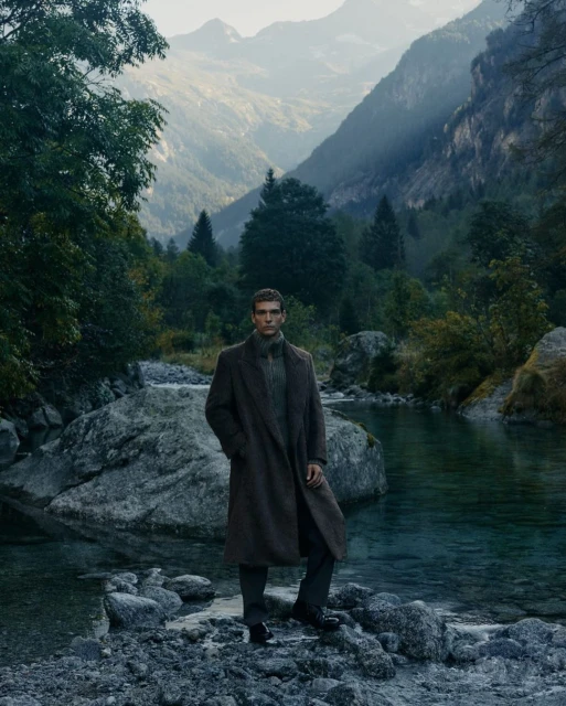 Man in coat stands on rocks in river surrounded by mountains and trees on a cloudy day in outdoor fashion editorial setting.