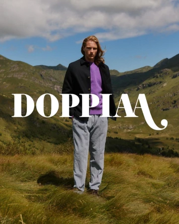 A model in outdoor fashion shoot, wearing a black jacket and grey pants, standing in a grassy field with mountains in the background.