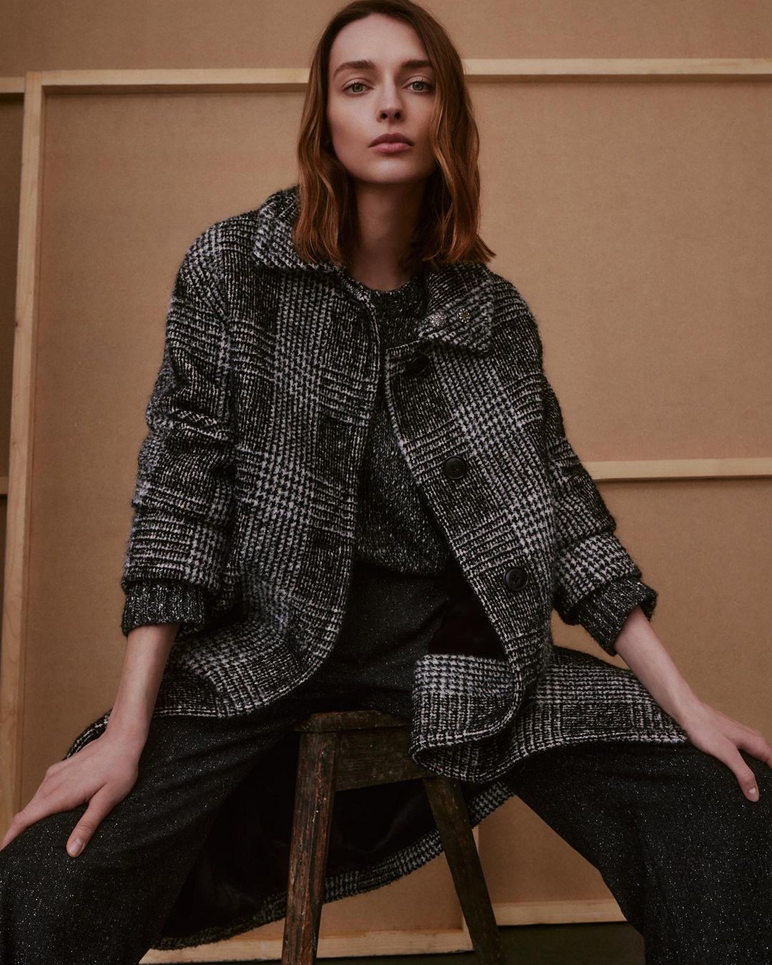 Woman wearing a black and white plaid jacket sitting on a wooden stool in front of a beige background in a fashion editorial image.