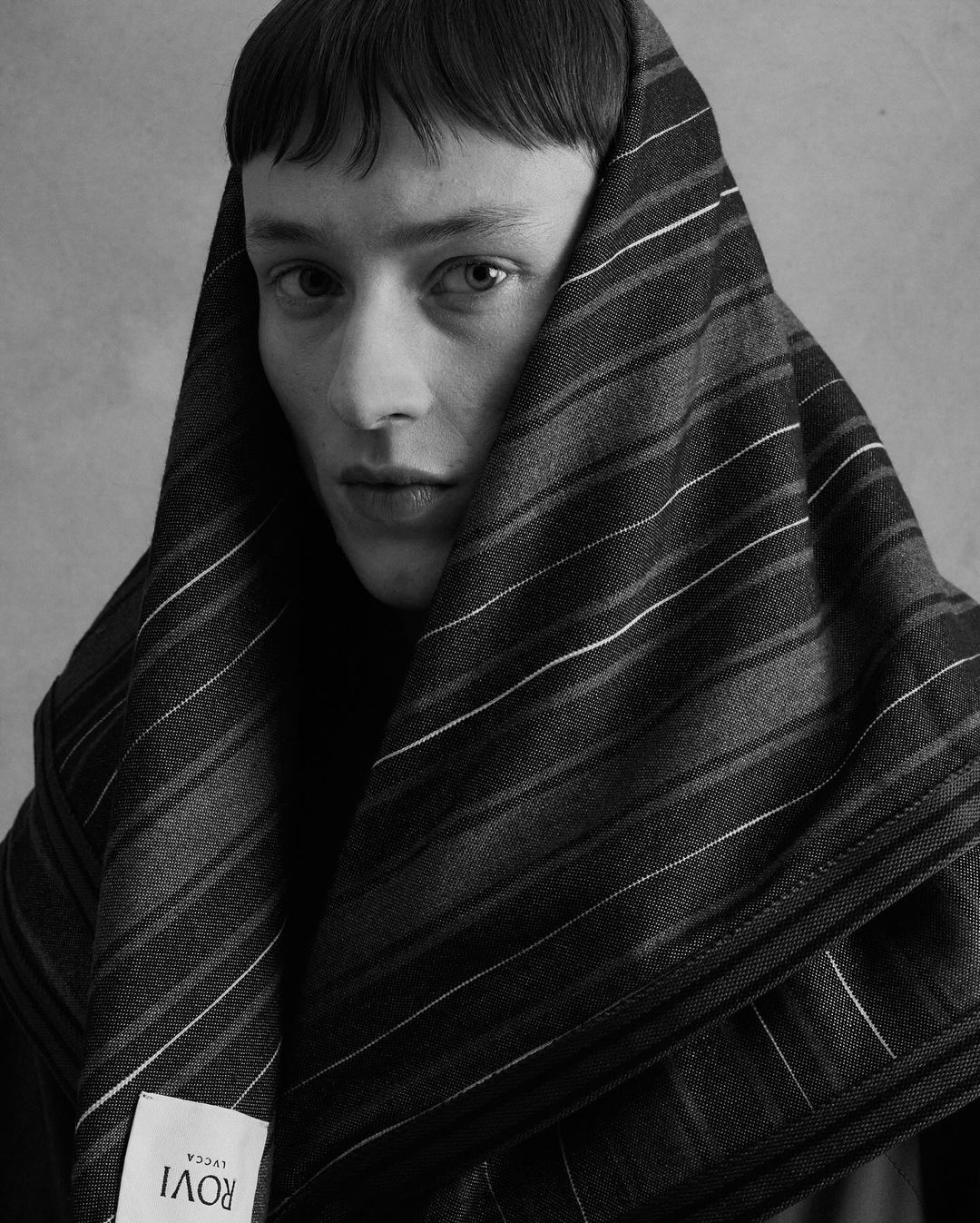 Monochrome portrait of a young adult with short hair, draped in a striped textile, gazing intently at the camera with soft lighting.