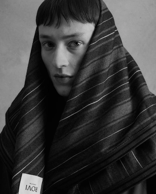 Monochrome portrait of a young adult with short hair, draped in a striped textile, gazing intently at the camera with soft lighting.