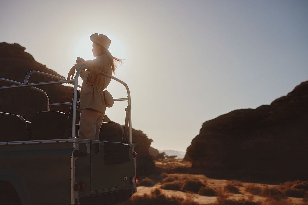 Woman in tan jacket and hat stands in an open jeep in a desert landscape at golden hour with rocky hills and a clear sky.