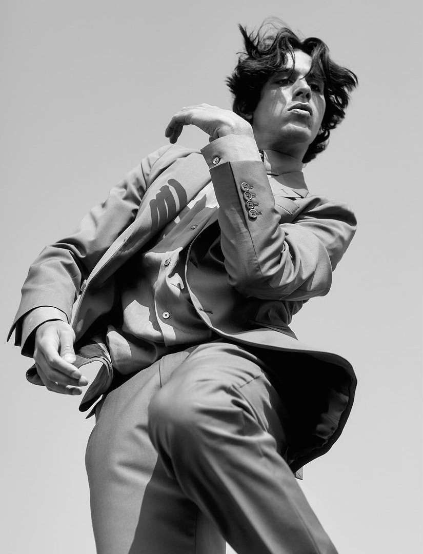 Young man in a suit poses against a blue sky in a black and white shot. He's wearing a button-down shirt and stylish suit.