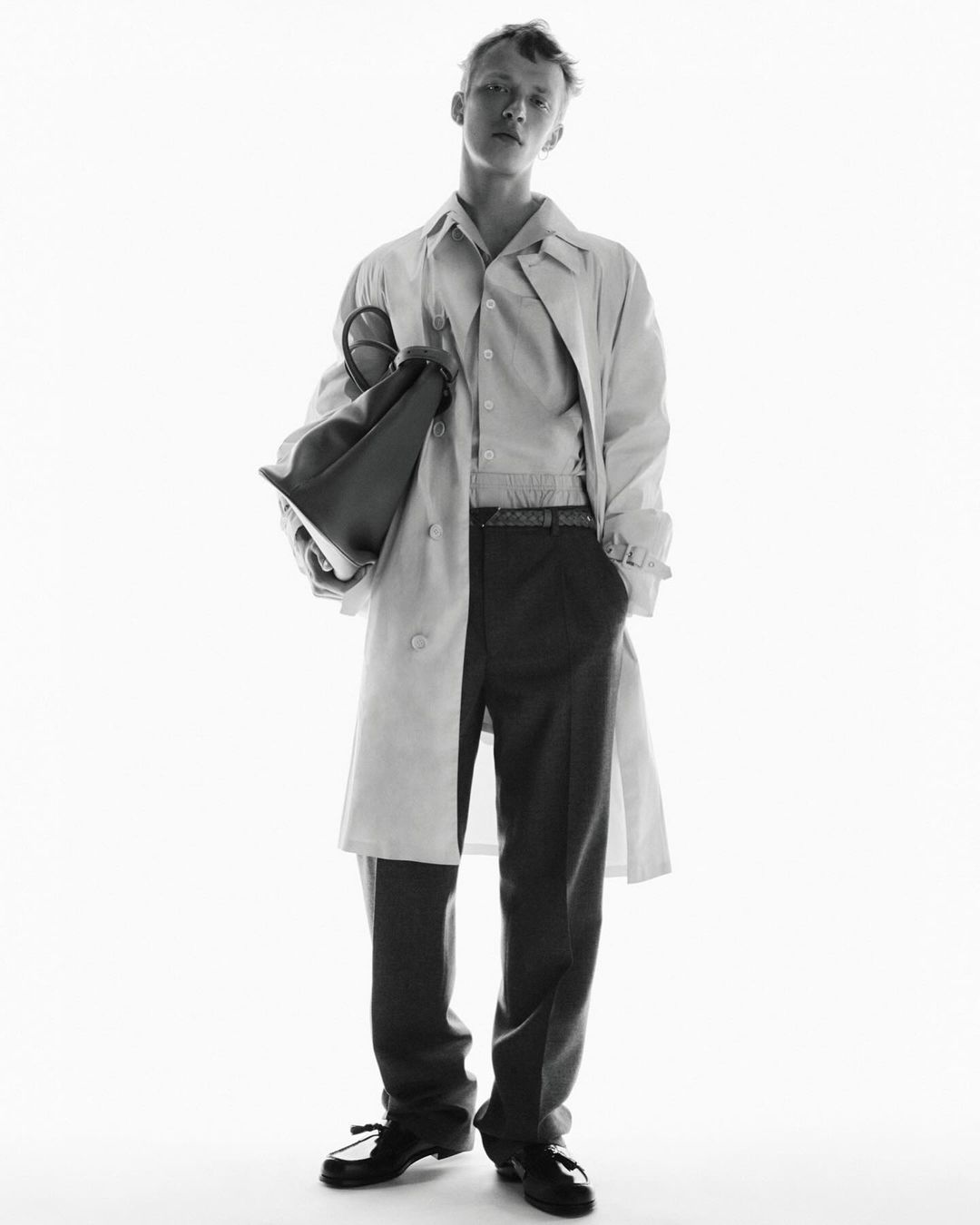 Black and white studio shot of a young adult man in a trench coat holding a bag, looking at the camera in a full shot.