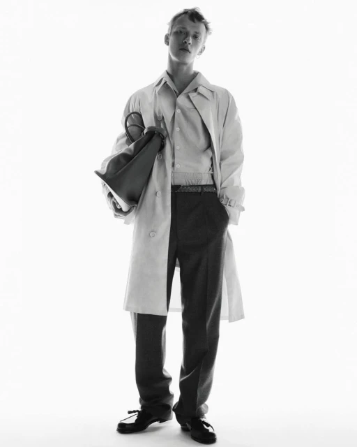 Black and white studio shot of a young adult man in a trench coat holding a bag, looking at the camera in a full shot.