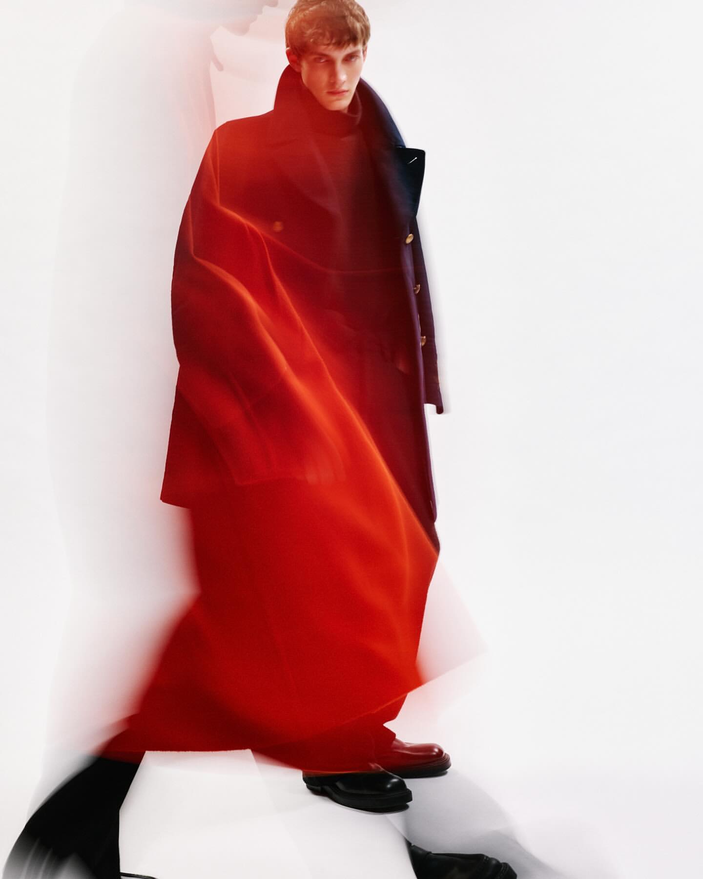 Fashionable adult male model in a long dark coat and glossy shoes, highlighted by red and white lighting in a neutral indoor setting.