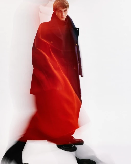 Fashionable adult male model in a long dark coat and glossy shoes, highlighted by red and white lighting in a neutral indoor setting.