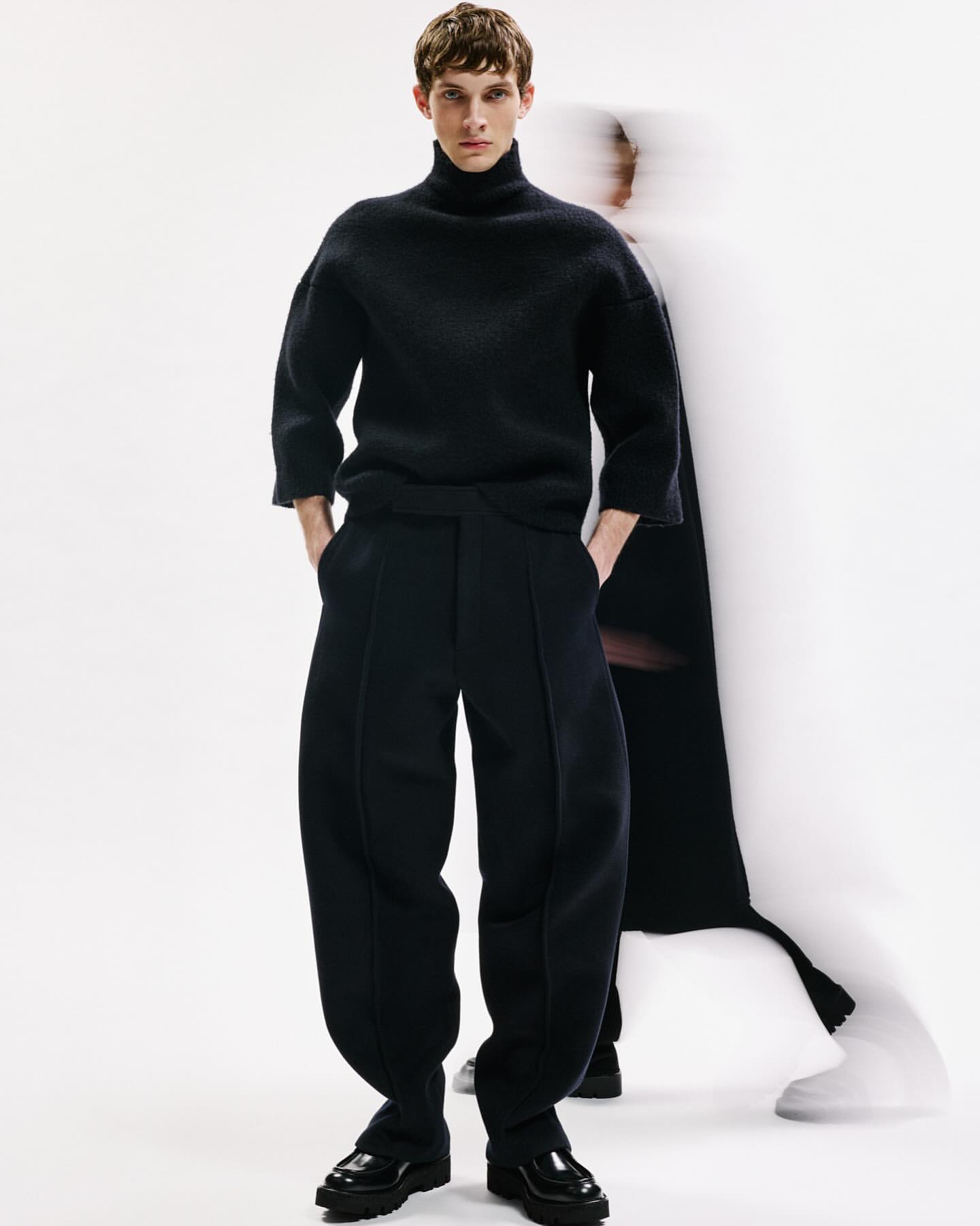A male model in a black turtleneck sweater and oversized trousers poses in a studio with a blurred figure in the background.
