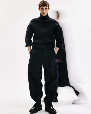 A male model in a black turtleneck sweater and oversized trousers poses in a studio with a blurred figure in the background.
