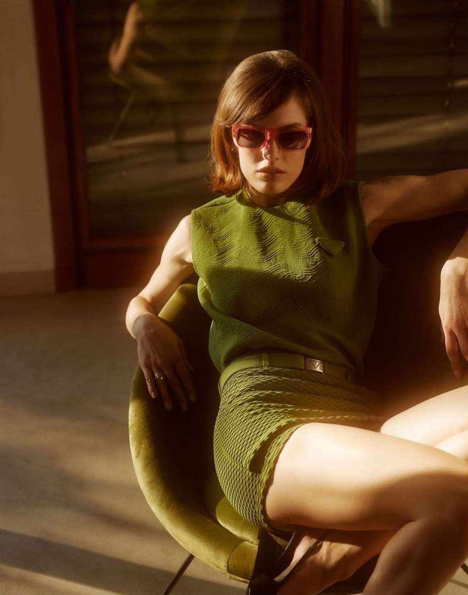 A woman in a green knit top and shorts poses in an olive-green chair, bathed in warm light indoors, wearing pink-framed sunglasses.