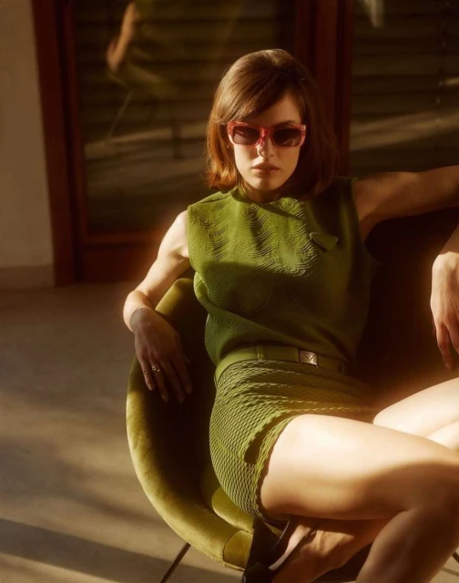 A woman in a green knit top and shorts poses in an olive-green chair, bathed in warm light indoors, wearing pink-framed sunglasses.