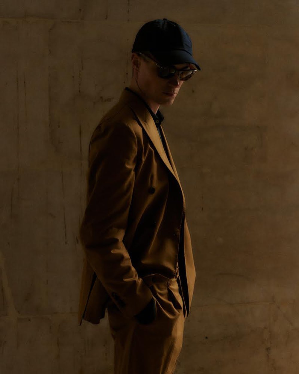 Man wearing a brown suit jacket, baseball cap, and sunglasses against a textured wall