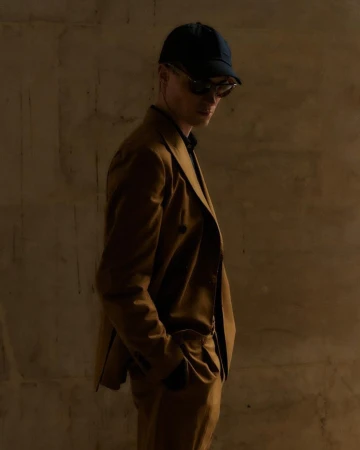 Man wearing a brown suit jacket, baseball cap, and sunglasses against a textured wall