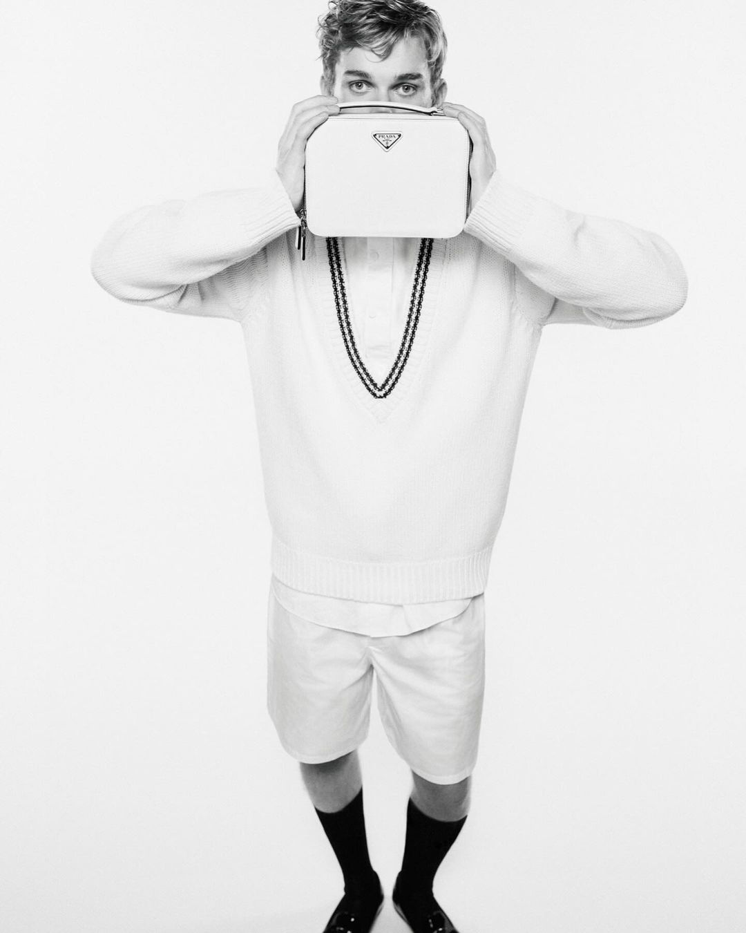 Black and white shot of a young adult man in a white sweater and shorts holding a white Prada handbag in front of his face.