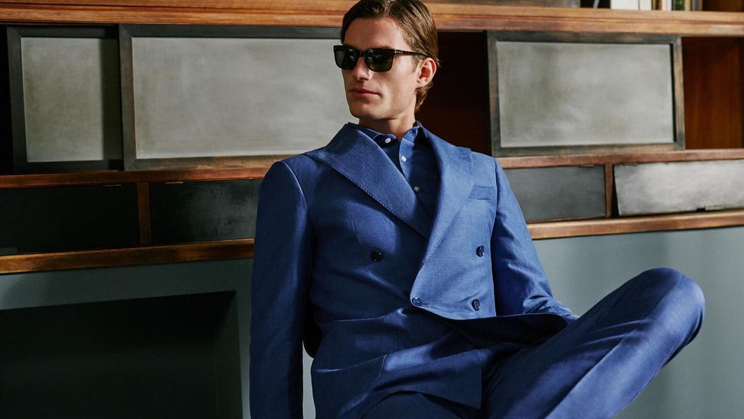 Man wearing a blue double-breasted suit and sunglasses seated against a wooden shelf background