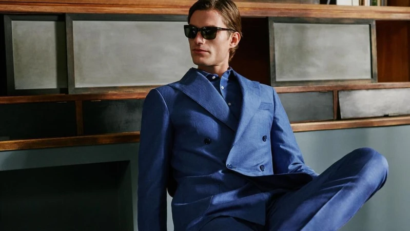 Man wearing a blue double-breasted suit and sunglasses seated against a wooden shelf background