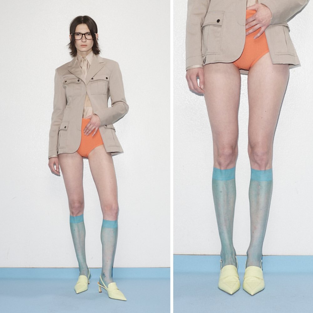 Woman in tan jacket and orange shorts posing in front of a white wall with blue trim on the bottom. She is also wearing light blue socks.