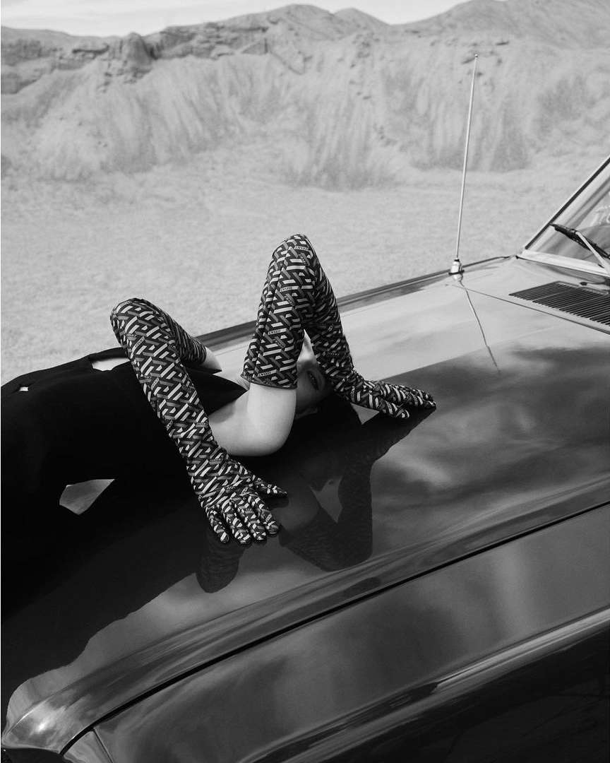 Black and white fashion shot of a person lying on the hood of a car in the desert, wearing gloves and thigh-high boots with a patterned design.