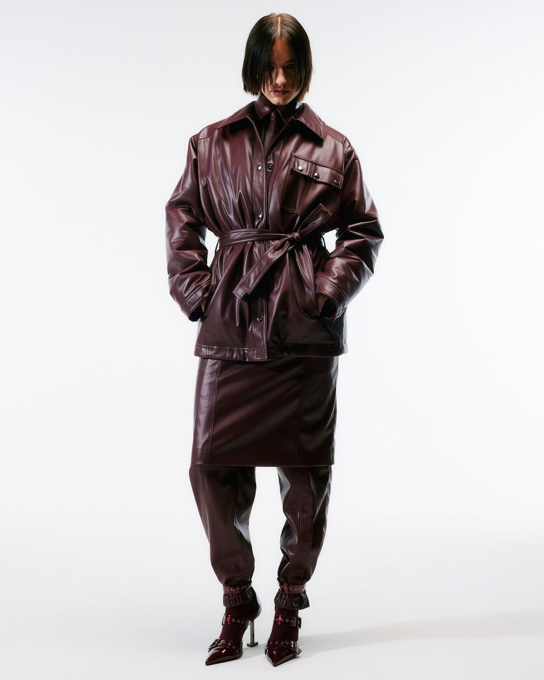 A woman in a burgundy leather jacket, skirt, and boots stands in a studio setting. The outfit is cinched at the waist with a belt.