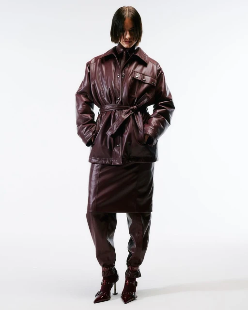A woman in a burgundy leather jacket, skirt, and boots stands in a studio setting. The outfit is cinched at the waist with a belt.