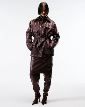 A woman in a burgundy leather jacket, skirt, and boots stands in a studio setting. The outfit is cinched at the waist with a belt.