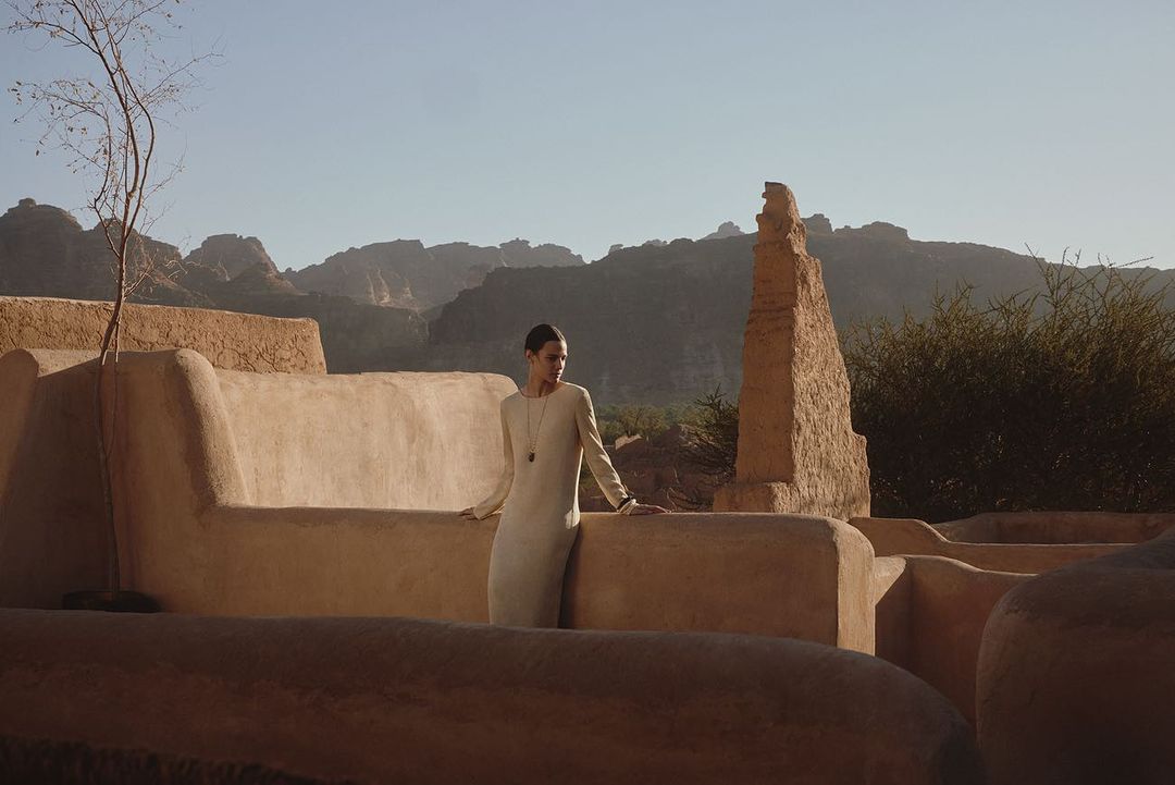 A woman in a long dress stands on a stone terrace with mountains in the background, showcasing a blend of fashion and landscape photography.
