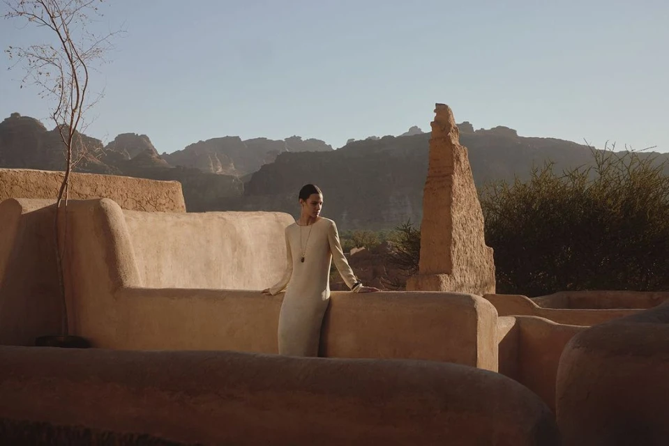 A woman in a long dress stands on a stone terrace with mountains in the background, showcasing a blend of fashion and landscape photography.