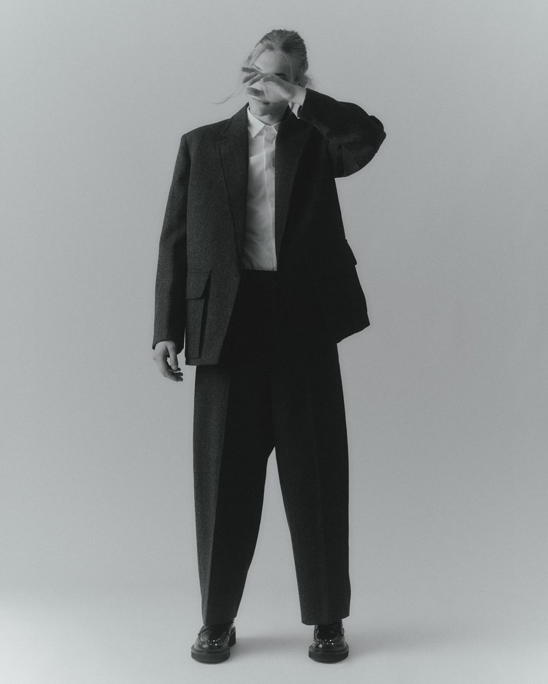 Monochrome studio shot of a person in an oversized blazer, dress shirt, and trousers with their hand covering their face.