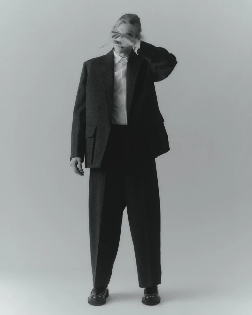 Monochrome studio shot of a person in an oversized blazer, dress shirt, and trousers with their hand covering their face.