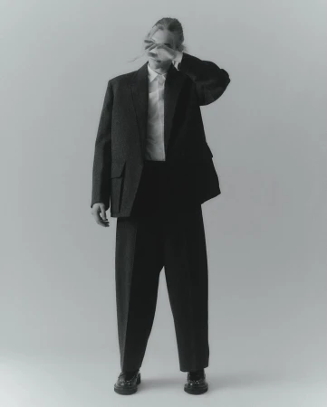 Monochrome studio shot of a person in an oversized blazer, dress shirt, and trousers with their hand covering their face.