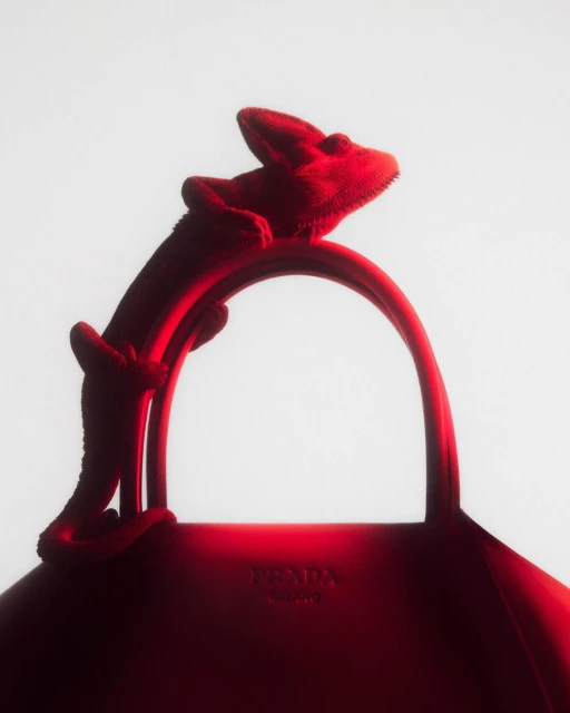 A vibrant red Prada handbag is prominently featured, artfully accented by a chameleon perched on its handle, against a bright background.