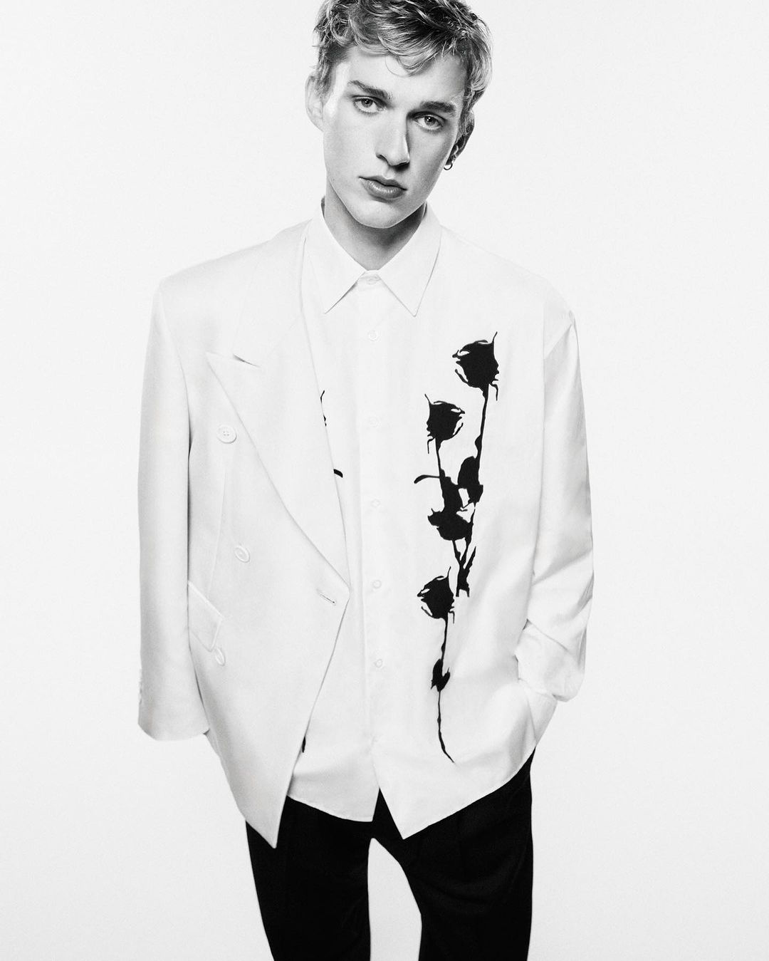 Fashion portrait of a young adult man in a white suit jacket with a black floral design on a white background.