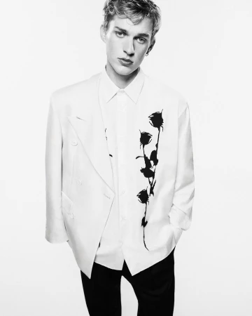 Fashion portrait of a young adult man in a white suit jacket with a black floral design on a white background.
