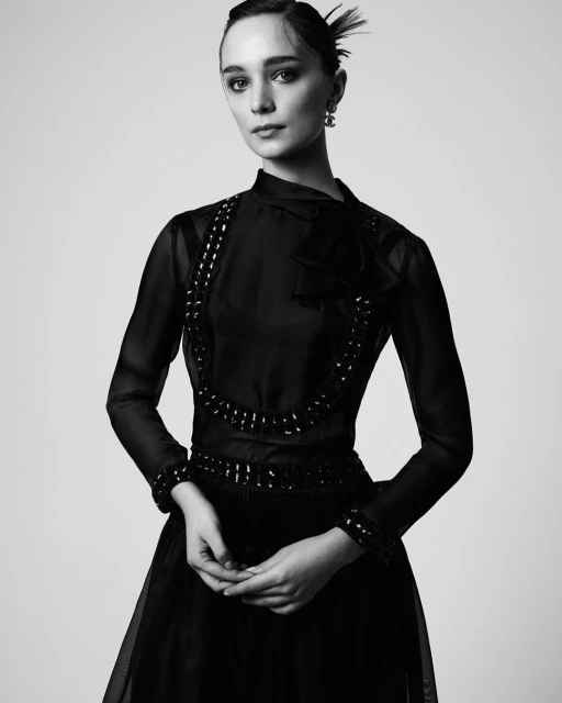 Black and white portrait of a woman in a black, sheer, long-sleeved dress with embellishments, posing against a neutral background.