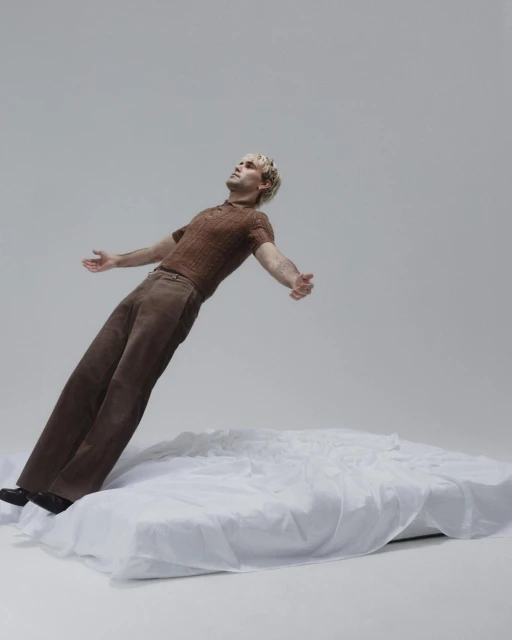 Adult male in brown knit shirt and trousers reclining on white fabric against a gray backdrop in a studio setting with soft lighting.