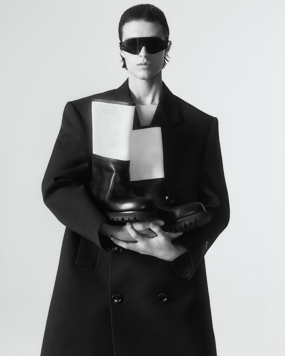 Fashion shot of a person in a black coat holding black and white boots with sunglasses against a white background in a photo studio.