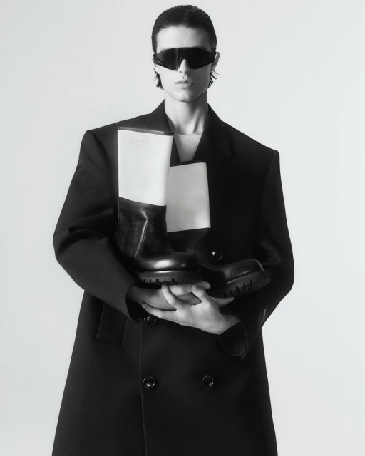 Fashion shot of a person in a black coat holding black and white boots with sunglasses against a white background in a photo studio.