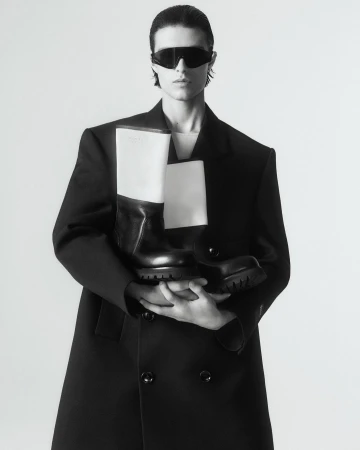 Fashion shot of a person in a black coat holding black and white boots with sunglasses against a white background in a photo studio.