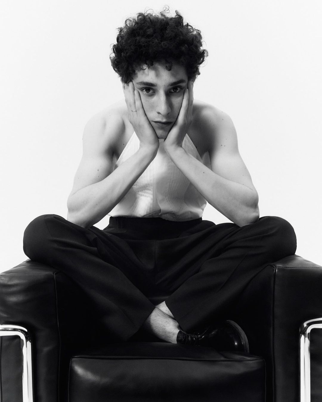 Black and white portrait of a young adult male with curly hair sitting cross-legged in a black leather chair in a bright studio.