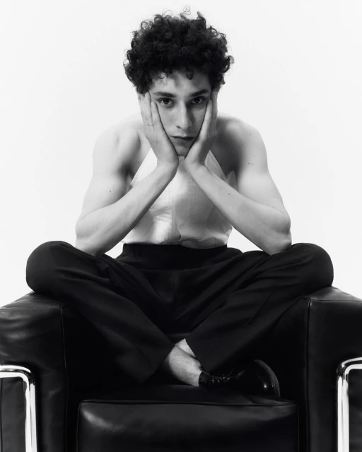 Black and white portrait of a young adult male with curly hair sitting cross-legged in a black leather chair in a bright studio.