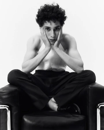 Black and white portrait of a young adult male with curly hair sitting cross-legged in a black leather chair in a bright studio.