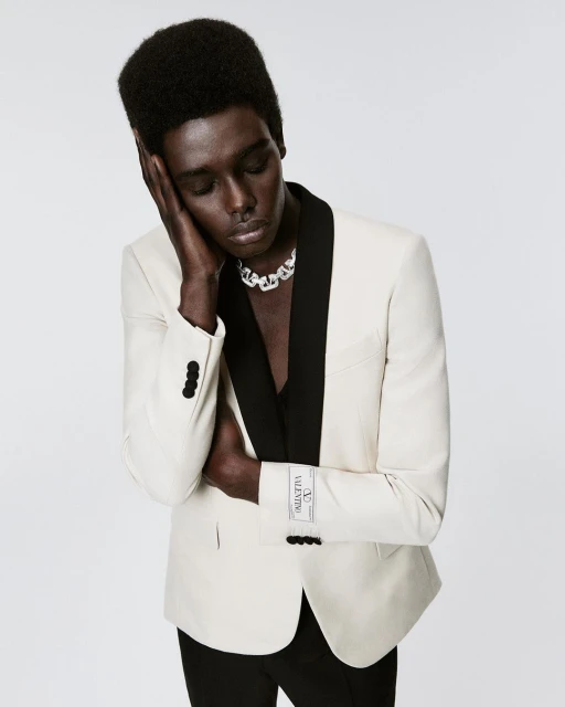 Man in white suit jacket with black lapel and chain necklace poses in a studio, looking down with hand against head.