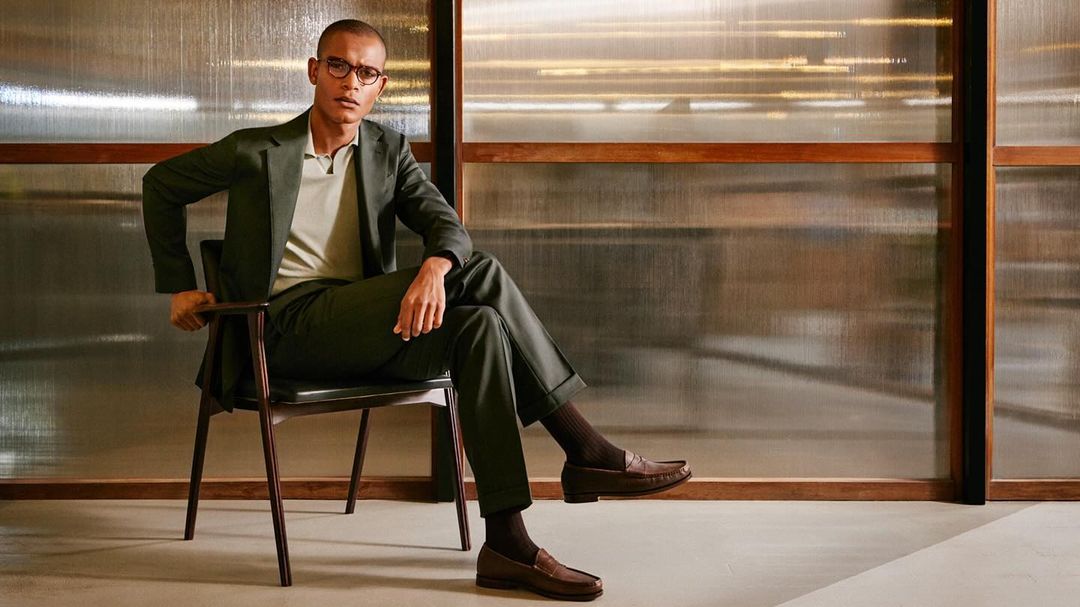 A sophisticated adult male in a green suit, eyeglasses, and brown loafers sits on a chair indoors, exuding a composed and elegant style.